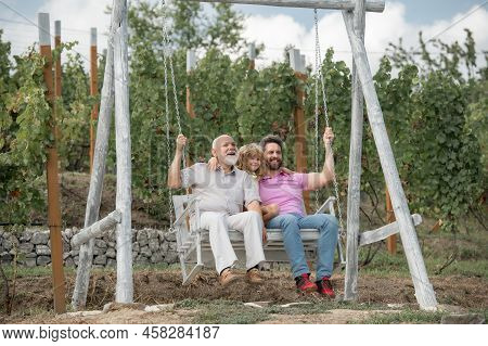 Boy Son With Father And Grandfather Swinging Together In Park Outdoors. Three Different Generations 