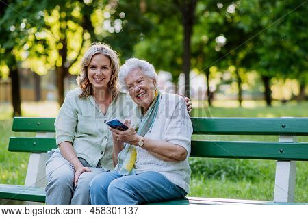 Adult Granddaguhter Helping Her Grandmother To Use Cellphone When Sitting On Bench In Park In Summer