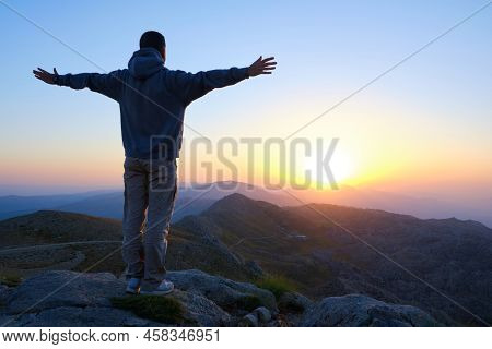Male Stands Towards The Sun On Mountain Slope With Spread Arms. Man Meets The Dawn With Open Arms. S