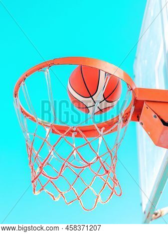 An Orange Basketball Ball In Hoop On A Blue Sky Background. A Ball Flying Into The Basket. Side Bott