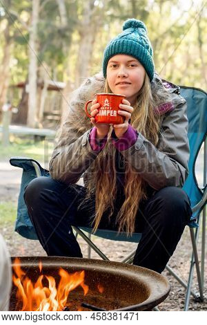 Camping Lifestyle Concept. Teenage Girl Wearing Beanie Hat Sitting In A Camping Chair Near Campfire