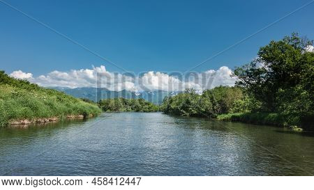 The Blue River Flows Calmly. There Is Lush Green Vegetation On The Banks - Grass, Trees. A Picturesq