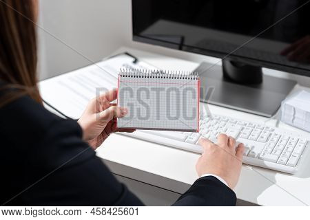Businesswoman Holding Important Message Wtitten On Notebook On Desk With Computer, Clipboard And Not