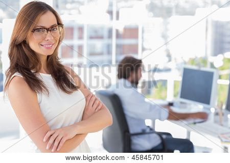 Sorrindo designer em pé em seu escritório com braços cruzados e colega atrás no trabalho