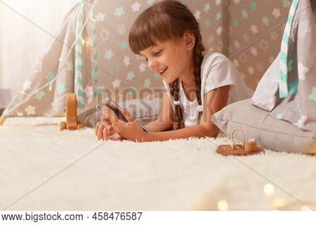 Side View Portrait Of Cute Little Girl With Pigtails Wearing White T Shirt Posing In Wigwam At Home 