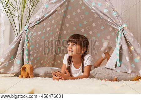 Horizontal Shot Of Dreamy Little Girl With Pigtails Wearing White T Shirt Posing In Wigwam At Home, 