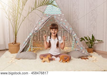 Indoor Shot Of Excited Extremely Happy Dark Haired Little Girl Sitting On Floor On Soft Carpet And C