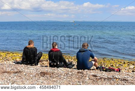 Three People Sit On The Seashore In Diving Clothes And Look Into The Distance.