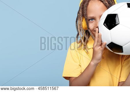 Smiling Little Boy With African Dreads Cover Face With Soccer Ball Over Blue Background.