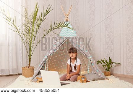 Indoor Shot Of Dark Haired Little Girl Sitting In Wigwam And Using Laptop Having Happy Facial Expres