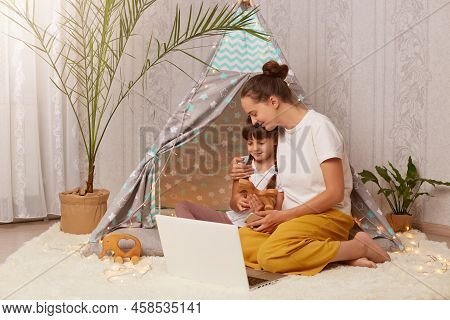Horizontal Shot Of Lovely Woman And Her Daughter Wearing White T Shirts Sitting In Wigwam In Front O