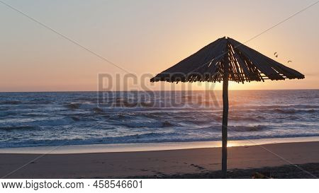 Beach Ocean Shoreline Man  Silhouetted Sitting Next To Wood Umbrella Watching Sunrise On The Sea Hor