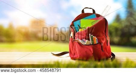 Backpack Full Of School Supplies On A Wooden Bench Outside With Gardens And School In The Background