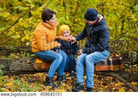 Mother, Father And Little Son Sit On A Plaid On A Log In The Autumn Park And Spend Time Together. Ho
