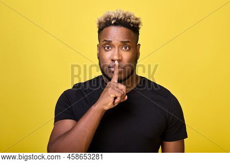 Serious Black Man Put His Index Finger To His Mouth Calling For Silence, Against A Yellow Background