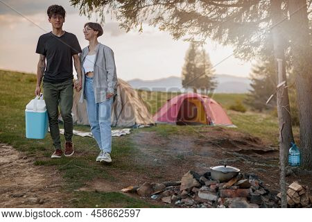 Young Man And Woman Standing Hand In Hand At Camping Place With Portative Fridge.