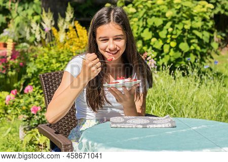 A Smiling Girl In The Garden At The Table Eats A Milk Strawberry Dessert.