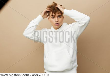 A Stylish Young Man In A White Hoodie Stands On A Beige Background And Pulls His Curly Hair Aside Wi