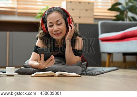 Joyful Middle Aged Woman Wearing Headphone, Using Moiled Phone While Lying On Floor At Home