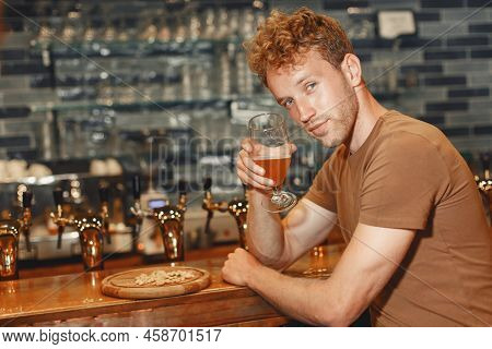 Red-haired Man Is Sitting At A Bar Drinking Beer From A Glass.