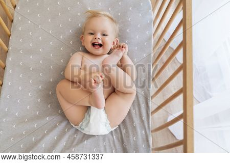 Upper View Portrait Of Funny Cute Baby In Diaper Lying On His Back In Bed Playing With Feet