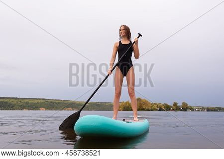 A Beautiful Girl Is Floating On A Supboard On A River. Young Woman Supsurfing At Sunrise.