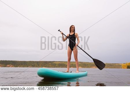 A Beautiful Girl Is Floating On A Supboard On A River. Young Woman Supsurfing At Sunrise.