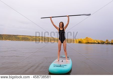 A Beautiful Girl Is Floating On A Supboard On A River. Young Woman Supsurfing At Sunrise.