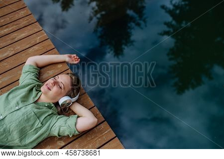 Rlaxed Woman Wearing Headphones Listening To Music Lying On A Pier By Natureal Lake In Summer