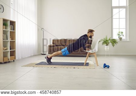 Athletic Man Is Doing Push Up Exercise With Chair At Home In His Living Room With Modern Interior.