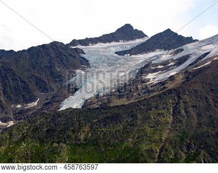 View Of Mount Cheget From The Village Of Terskol. A High Mountain With Snowy Peaks Against A Blue Sk