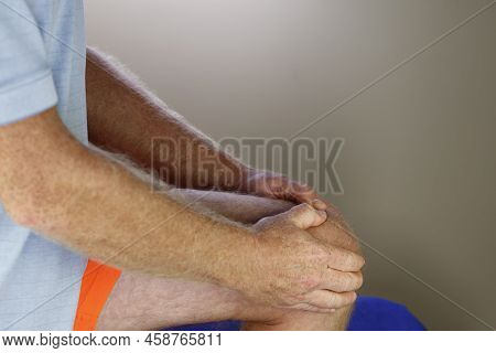 Left Knee Being Self Massaged By A Mature Caucasian Man Wearing Orange Sports Clothes Blue Shirt Clo