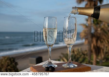 Pouring Of Spanish Cava Sparkling Wine Is Glasses With View On Blue Sea And Sandy Beach, Costa Del S