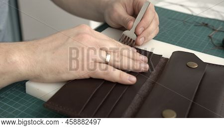 Mens Hand Holding A Hammer And Hole Punch And Makes A Holes For Seawing A Leather Wallet In His Work