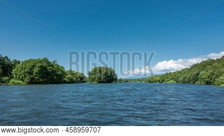 There Is Lush Green Vegetation On The Banks Of A Wide, Calm River. Cumulus Clouds In A Blue Sky. Cop