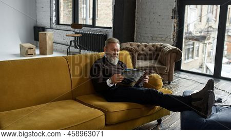 Grey Hair Concentrated Caucasian Man Reading Journal On Sofa At Home. Fashionable Bearded Pensioner.