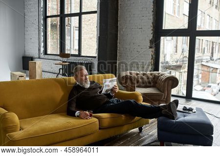 Senior Focused Caucasian Man Drinking Whiskey And Reading Newspaper On Sofa At Home. Stylish Bearded