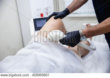 A Male Cosmetologist In A Dark Blue Uniform Conducts A Procedure Of Laser Hair Removal Of The Legs O