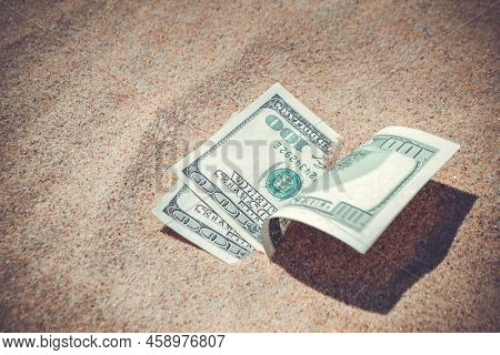 Money Dollars Half Covered With Sand Lie On Beach Close-up. Dollar Bills Partially Buried In Sand. T
