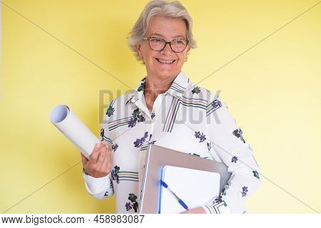 Confident Expert Architech Senior Woman With Laptop And Notebook In Hand, Isolated On Yellow Backgro