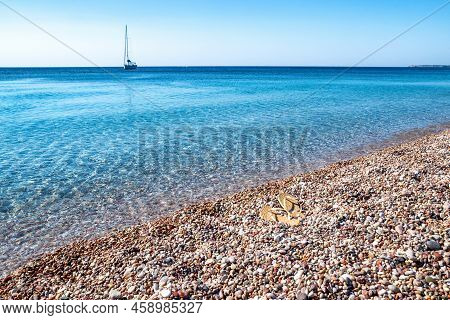 Sandals On The Shore With Pebble Beach. Yacht On The Sea At Background.