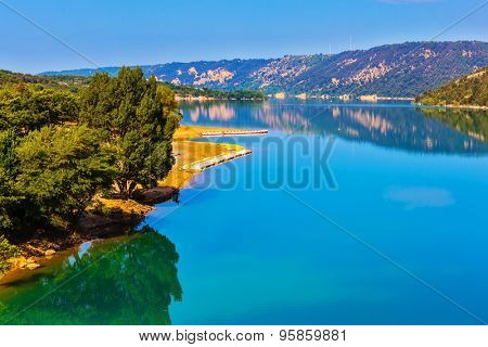 Smooth emerald green water of Lake Sainte-Croix-du-Verdon reflects the sky and wooded shore. Mountain canyon Verdon in the French Alps