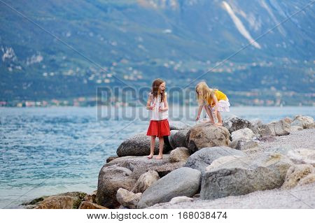 Two Cute Little Sisters Having Fun On A Beach Of Limone Sul Garda, A Small Town And Comune In The Pr