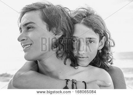 Greyscale portrait of young long haired man looking away and curly lady hugging him from the back standing at the beach