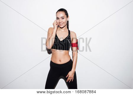 Happy charming young fitness woman with armband standing and listening to music over white background