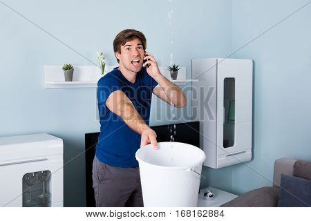 Worried Man Calling Plumber While Leakage Water Falling Into Bucket At Home