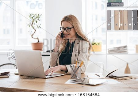 Important business talk. Confident young woman in smart casual wear talking on the mobile phone and smiling while sitting at her working place in creative office