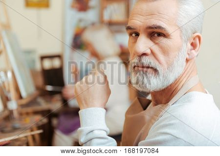 Think of art. Elderly concentrated handsome artist sitting in painting class and posing while enjoying his hobby.