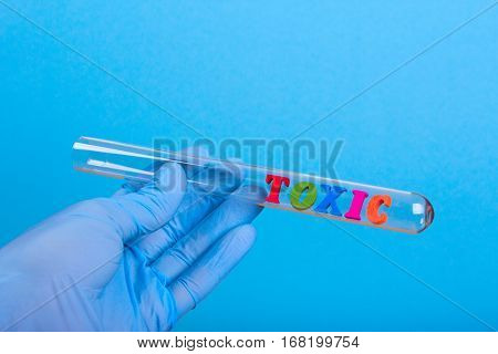 Word toxic of colored wooden letters in test-tube. Hand in glove holding a test tube on a blue background