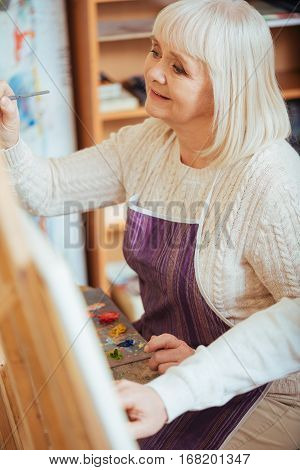 Make life art. Delighted elderly concentrated woman painting on a canvas while using brush and enjoying her hobby.